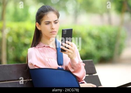 Una donna convalescente preoccupata che controlla il telefono seduto su una panchina in un parco Foto Stock