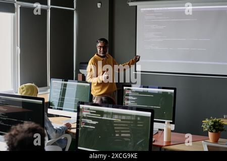 Allegro e maturo insegnante di sesso maschile in piedi di fronte agli studenti che spiega i problemi di codifica durante la lezione di informatica Foto Stock