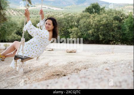 Una giovane donna con un abito estivo bianco oscilla gioiosamente in un ambiente panoramico all'aperto circondato da alberi e colline. Cattura l'essenza del lei spensierato Foto Stock