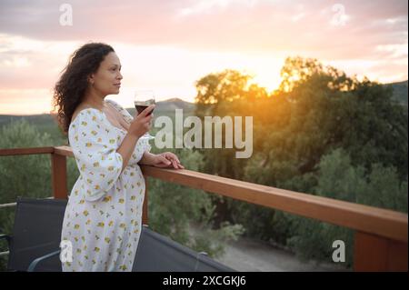 Una donna con un abito bianco floreale gode di un bicchiere di vino mentre guarda il tramonto da un balcone di legno che si affaccia su un paesaggio panoramico. Foto Stock