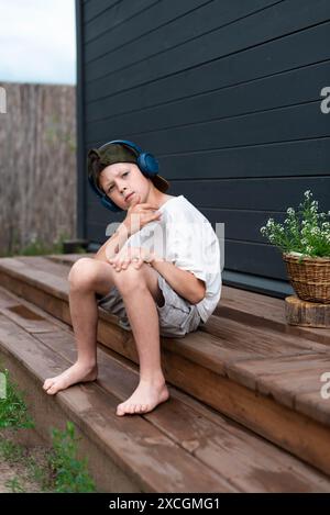 Simpatico ragazzo preadolescente con le cuffie che ascolta la musica su una terrazza in legno all'aperto Foto Stock