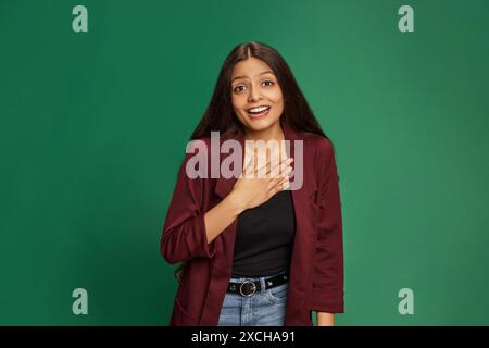 Ritratto di una giovane donna indiana con lunghi capelli scuri che si posa sorprendentemente alla fotocamera e tiene la mano sul petto sullo sfondo verde dello studio. Foto Stock