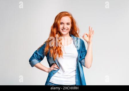Donna dai capelli rossi sorridente che dà il segno di Okay sul bianco Foto Stock