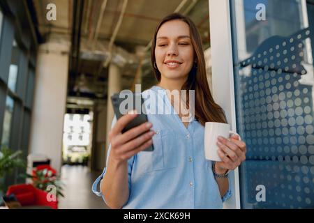 Una donna sembra felice di sfogliare il suo smartphone tenendo una tazza di caffè in un ufficio alla moda Foto Stock