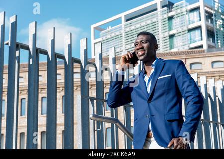Sorridente professionista afroamericano che cammina con lo smartphone tra i moderni uffici del centro città. Foto Stock