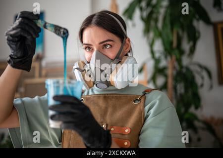 Donna concentrata e attenta nel respiratore, guanti che lavorano con resina epossidica, creando arti, dipinti. Foto Stock
