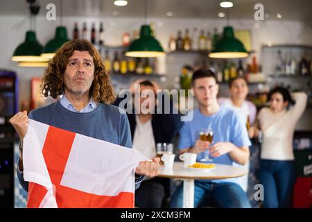 Compagnia di giovani appassionati di sport adulti turbati che detengono la bandiera inglese e si preoccupano della sconfitta durante la partita in TV nel pub Foto Stock