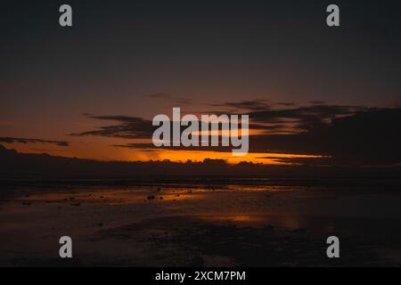 Un tramonto panoramico sulla spiaggia con spettacolari nuvole e riflessi sulla sabbia bagnata. I colori vivaci creano una scena mozzafiato e serena, che mette in mostra la bellezza naturale del paesaggio costiero. Foto Stock