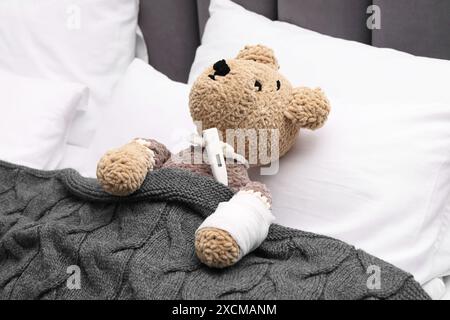 Simpatico orsacchiotto con bendatura e termometro sotto coperta nel letto Foto Stock