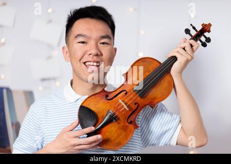 Un adolescente asiatico sorride allegramente mentre tiene un violino nella sua stanza a casa Foto Stock