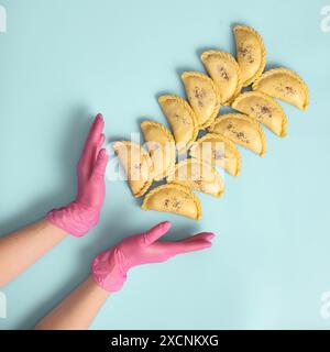 Flat poggia le mani di una donna fornaio con guanti di gomma rosa che staglia samsa crudo su sfondo blu. Cucina indiana tradizionale, congelata e semilavorata Foto Stock