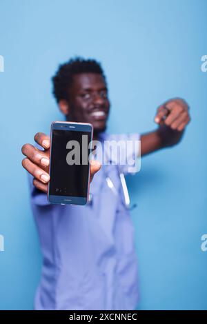 Il medico afroamericano in scrub tiene il dispositivo mobile con schermo vuoto. Primo piano di un infermiere medico con impugnature stetoscopio e gesti verso lo smartphone con display vuoto. Foto Stock