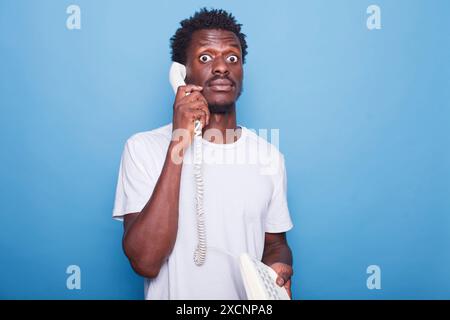 Uomo nero sorpreso con capelli afro su sfondo blu che parla su un telefono fisso. Ritratto di un uomo afroamericano scioccato che comunica attraverso il telefono. Foto Stock