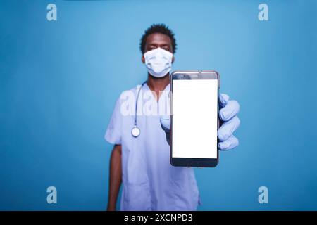 L'uomo afroamericano in scrub afferra un cellulare che mostra uno schermo bianco isolato. Primo piano di infermiere maschio che indossa la maschera, tenendo dispositivo mobile con modello cromakey bianco mockup. Foto Stock