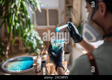 Artista donna seria nel respiratore che lavora professionalmente con resina epossidica, creando dipinti. Foto Stock