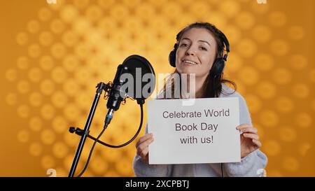 Donna sorridente che filma un video promozionale per la giornata mondiale del libro, isolato sullo sfondo dello studio. Influencer che promuove la lettura, sensibilizzazione per l'evento internazionale globale, camera B Foto Stock