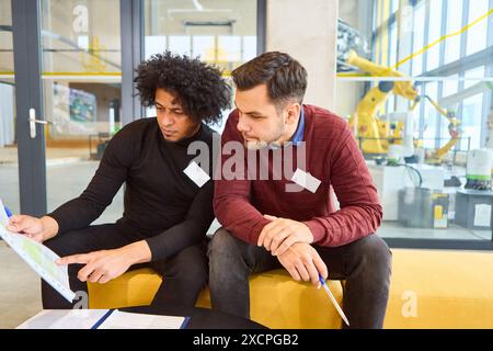 Due ingegneri analizzano i documenti e discutono il processo di programmazione e ispezione dei bracci robotici in una struttura industriale all'avanguardia. Foto Stock