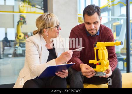 Due ingegneri discutono dello sviluppo di un braccio di presa robotizzato in un ambiente industriale moderno, concentrandosi sull'automazione e la programmazione. Foto Stock