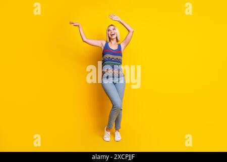 Ritratto fotografico di una giovane donna bionda energica in jeans e di un singlet che danzano al night club dancehall isolato su sfondo giallo Foto Stock