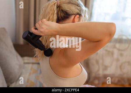 Ragazza a casa con una pistola massaggiante a percussione terapeutica in mano e massaggiando i muscoli della schiena concetto di recupero sportivo dopo un allenamento Foto Stock