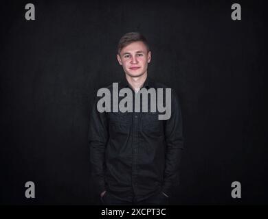 Ritratto di un giovane uomo sicuro e rilassato, tiene le mani in tasca su uno sfondo nero. Studente maschile spensierato con espressione facciale positiva Foto Stock