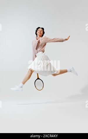 Immagine verticale a tutta lunghezza di una bella giovane donna in gonna e camicetta, in posa a mezz'aria con racchetta da tennis, che gioca isolata su sfondo bianco. Foto Stock