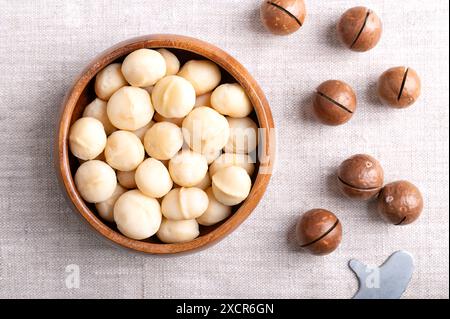 Noci di macadamia tostate in un recipiente di legno su tessuto di lino. Sulla frutta giusta con conchiglie tagliate e chiave di apertura. Foto Stock