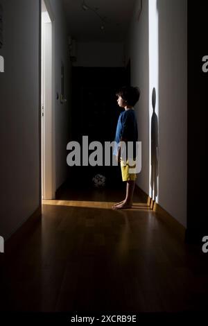 Un bambino si trova in un corridoio buio, la sua silhouette è stata proiettata a lungo sulla parete da un frammento di luce proveniente da una porta aperta. Foto Stock