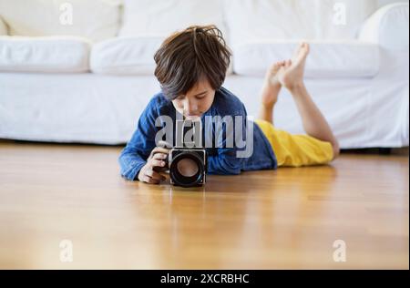 Un ragazzino giace sul pavimento in legno duro, guardando con attenzione attraverso l'obiettivo di una macchina fotografica vintage. Sta catturando il momento, concentrandosi sul trovare il perf Foto Stock