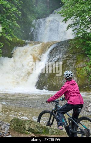 Donna anziana, in sella alla sua mountain bike elettrica e guardando la cascata di Eibele in alto mare nelle Alpi Allgau vicino a Oberstaufen, Baviera, Gemania Foto Stock