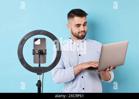 Blogger di tecnologia che esamina il laptop e registra video con smartphone e lampada anulare su sfondo azzurro Foto Stock