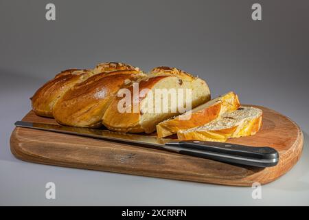 Un pane intrecciato appena sfornato, ricoperto di mandorle e ripieno di uvetta, è parzialmente tagliato a fette e esposto su un tagliere di legno. Il brea Foto Stock