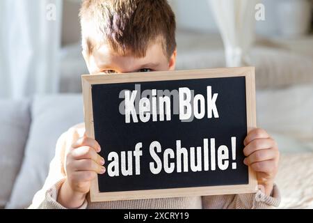 Germania - 18 giugno 2024: Un ragazzo ha un cartello con la lavagna su cui è scritto in gesso bianco Kein Bok auf Schulle , un'errata ortografia dovrebbe significare: Kein Bock auf Schule immagine simbolica di cadute scolastiche. FOTOMONTAGGIO *** Ein Junge hält ein Tafelschild, auf dem in Weißer Kreide Kein Bok auf Schulle geschrieben steht, Rechtschreibfehler soll heißen: Kein Bock auf Schule Symbolbild Schulabbrecher. FOTOMONTAGE Foto Stock