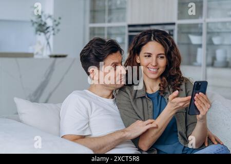La coppia si siede sul divano, rilassata e felice, guardando lo smartphone insieme. La donna sorride e mostra qualcosa al telefono all'uomo, che si appoggia e ascolta con attenzione. Foto Stock