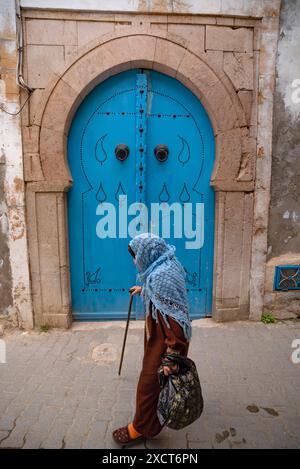 Tunisi, Tunisia. 7 maggio 2024. Una vecchia donna che indossa uno scialle blu passa davanti a una tradizionale porta blu in uno dei tanti vicoli stretti che compongono la Medina di Tunisi, una città patrimonio dell'umanità dell'UNESCO (Organizzazione delle Nazioni Unite per l'educazione, la scienza e la cultura), la Tunisia. (Foto di John Wreford/SOPA Images/Sipa USA) credito: SIPA USA/Alamy Live News Foto Stock