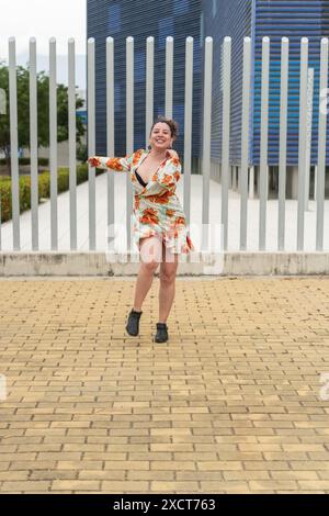 Donna che balla allegramente all'aperto con un abito floreale vicino a una recinzione di metallo e a un edificio moderno. Foto Stock