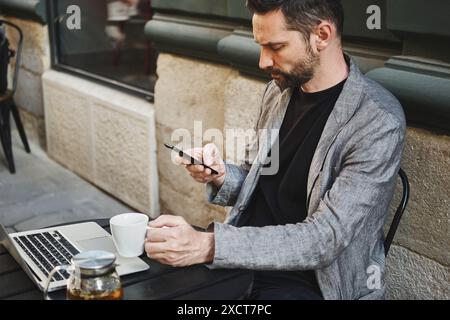 Un uomo caucasico si siede a un tavolo in un bar di strada e guarda il telefono Foto Stock