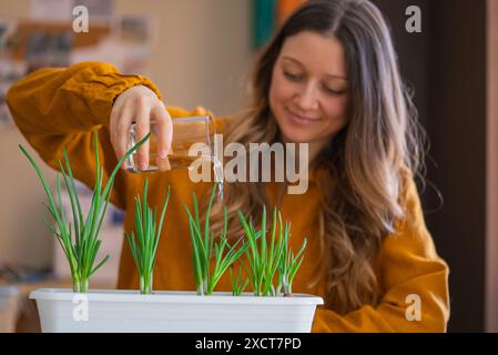 La donna che indossa un maglione color senape innaffia felicemente le piante di cipolla verde che crescono nella piantatrice bianca. Coltivare verdure per insalata a casa. Foto Stock