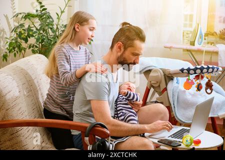 Padre caucasico lavora in multitasking mentre lavora su un laptop, tiene in braccio un bambino e viene assistito dalla figlia maggiore in un ambiente accogliente. Concetto di ufficio domestico Foto Stock