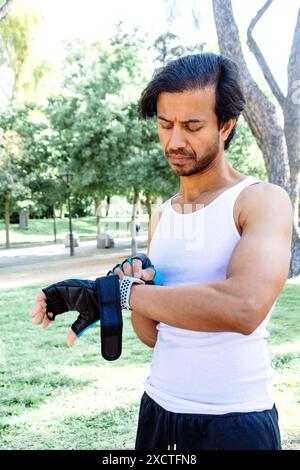 Un uomo con canotta bianca e pantaloncini neri indossa un cinturino da polso blu. E' in piedi in un parco ed e' concentrato su qualcosa Foto Stock