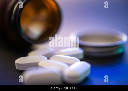 Primo piano di pillole bianche sparse vicino a un flacone di vetro ambrato aperto su una superficie blu scuro, concentratevi sulle pillole in primo piano, con sfondo sfocato. Foto Stock