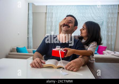 La figlia che chiude gli occhi di suo padre mentre legge un libro e gli dà una confezione regalo rossa Foto Stock