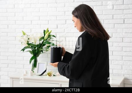 Giovane donna che mette l'urna mortuaria sul cassettiere vicino al bouquet di fiori di giglio funebre in camera Foto Stock