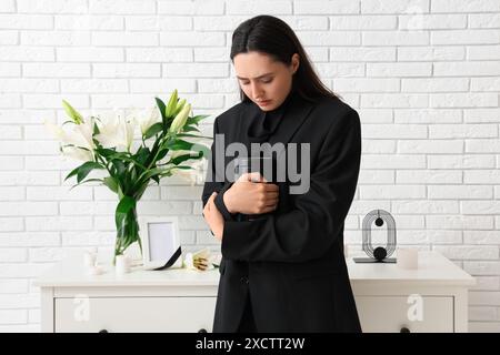 Giovane donna che tiene in mano urna mortuaria vicino al bouquet di fiori di giglio funebre in camera Foto Stock