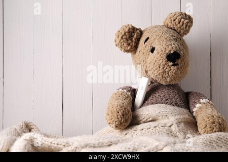 Orsetto giocattolo con termometro sotto coperta vicino a una parete di legno bianca, spazio per il testo Foto Stock