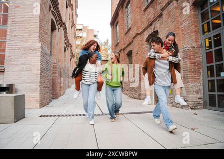 Gruppo di studenti maschi che fanno un giro in piggyback alle sue amiche, camminano o corrono fino alla classe. Eccitati compagni di classe multirazziali che si divertono insieme al campus universitario. Torna a scuola. Foto di alta qualità Foto Stock