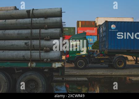 Giacarta, Indonesia - 9 maggio 2024. Un grande camion verde con un rimorchio giallo caricato con tubi di cemento è parcheggiato accanto a un contenitore blu Foto Stock