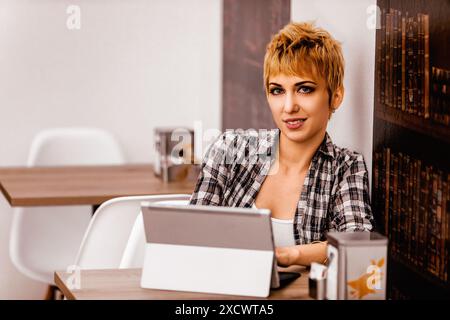 Giovane donna sicura di sé con i capelli biondi corti lavora su un laptop in un accogliente bar, incarnando il moderno stile di vita nomade digitale Foto Stock