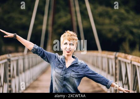 Giovane donna con i capelli biondi corti si erge su un ponte, braccia allungate, sorridenti e spensierati, godendosi la natura al sole. La sua camicia in denim reflec Foto Stock