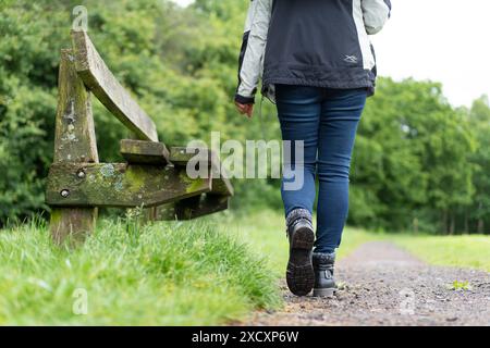 Punto di vista posteriore basso di una donna in jeans e stivali che cammina in un parco del Regno Unito accanto a una panchina di un parco su una pista di ghiaia. Foto Stock